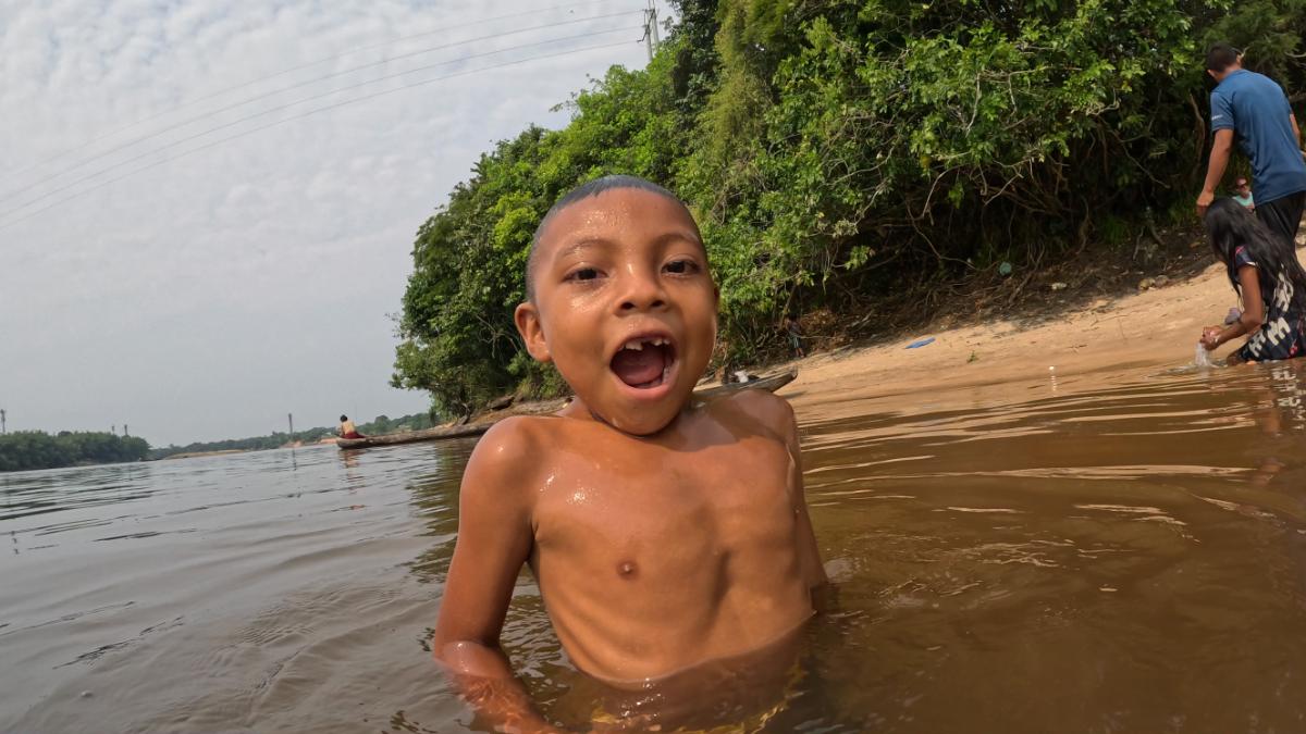 a young child wading and playing in a river, shot at water level with a sandy riverbank and other people visible in the background