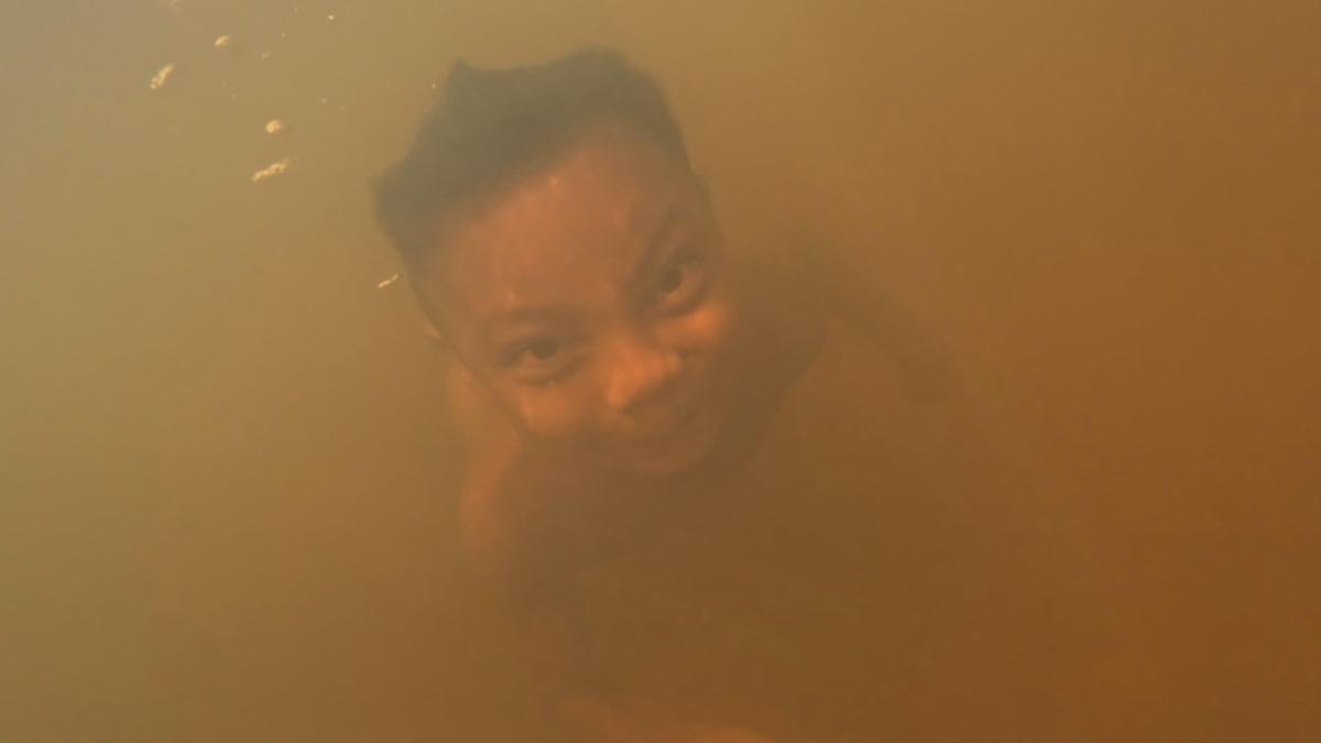 close-up of a person submerged in turbid amber-coloured water, face barely visible through the murk