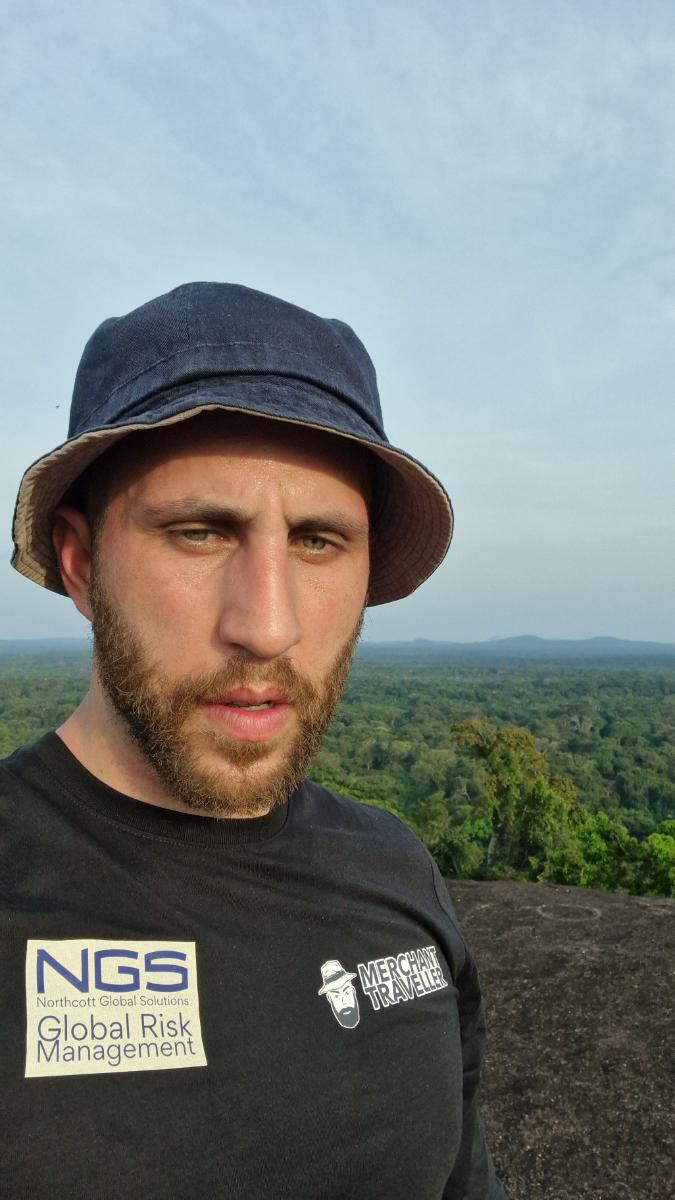 a man wearing an NGS Global Risk Management branded T-shirt at an elevated outdoor viewpoint with a forest canopy in the background