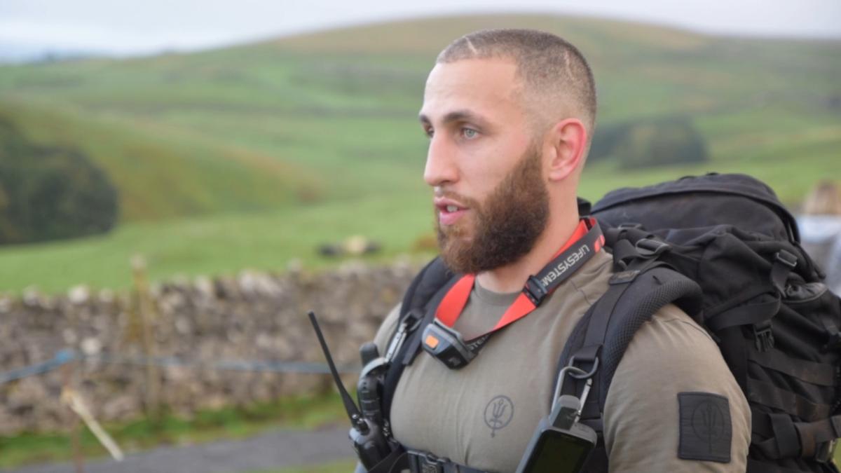 close-up portrait of a man in tactical gear with a large backpack and radio, speaking outdoors against a rural hillside