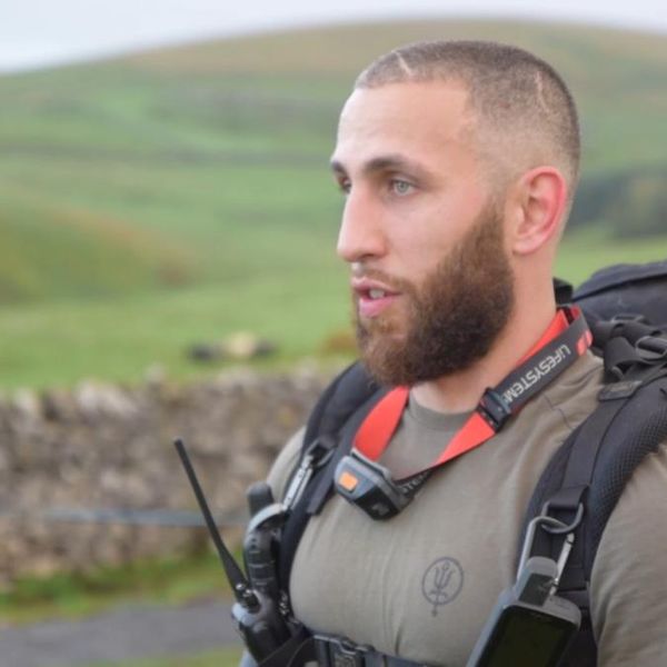close-up portrait of a man in tactical gear with a large backpack and radio, speaking outdoors against a rural hillside