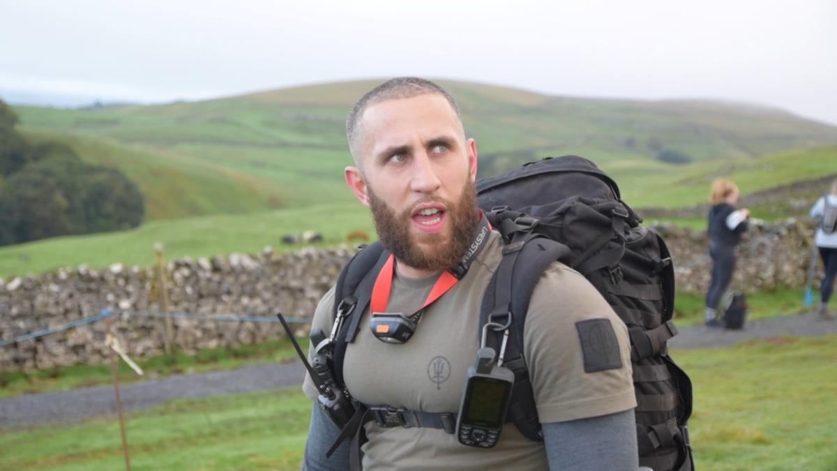 a man with a heavy tactical rucksack and GPS device pausing on a hillside trail