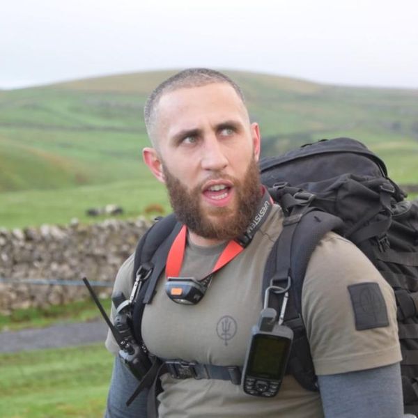 a man with a heavy tactical rucksack and GPS device pausing on a hillside trail