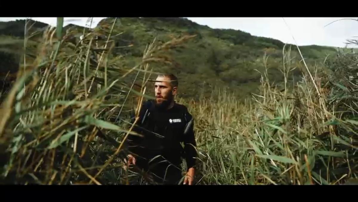 a bearded man in a black branded jacket moving through tall reeds or wild grass on a hillside with green mountains behind