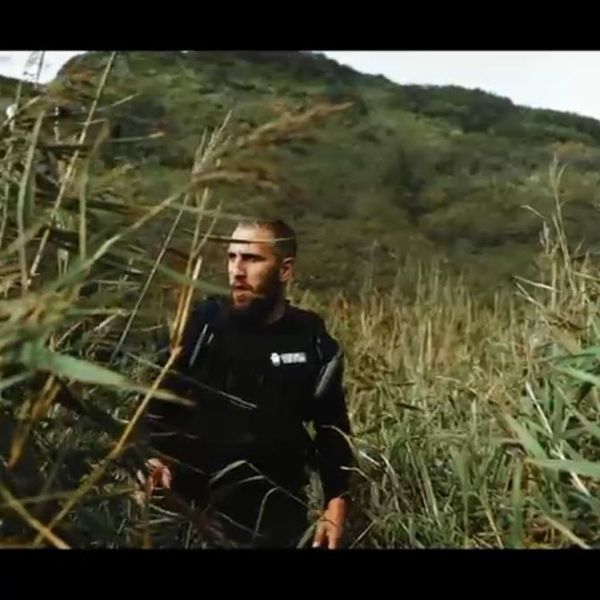 a bearded man in a black branded jacket moving through tall reeds or wild grass on a hillside with green mountains behind