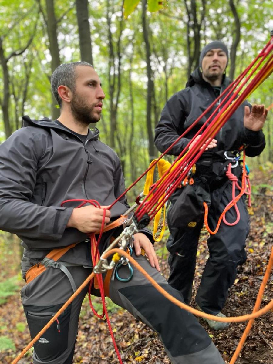 two people in climbing harnesses managing ropes and rigging equipment in a woodland setting