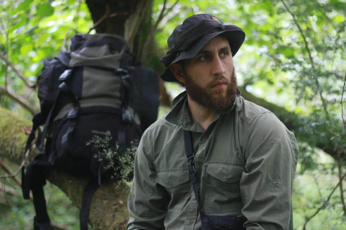 a man in outdoor field clothing pausing in woodland with a large backpack nearby