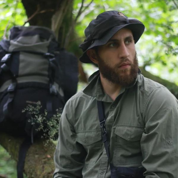 a man in outdoor field clothing pausing in woodland with a large backpack nearby