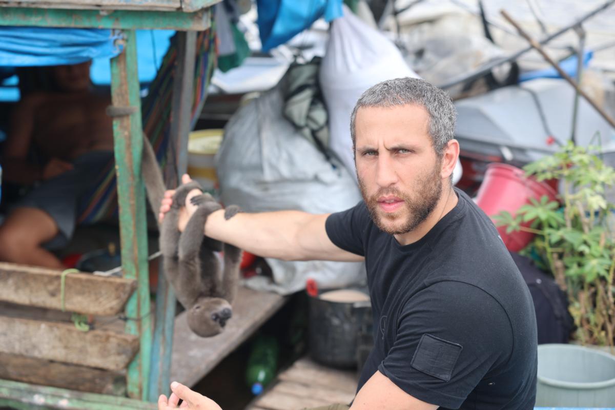 a man holding a small wild animal on a rustic boat, surrounded by sacks and supplies