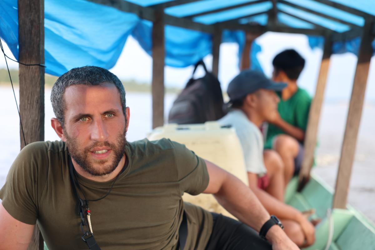 a man seated on a covered wooden river boat with other passengers, open water visible in the background