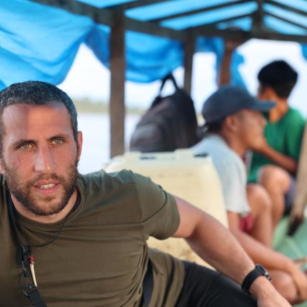 a man seated on a covered wooden river boat with other passengers, open water visible in the background