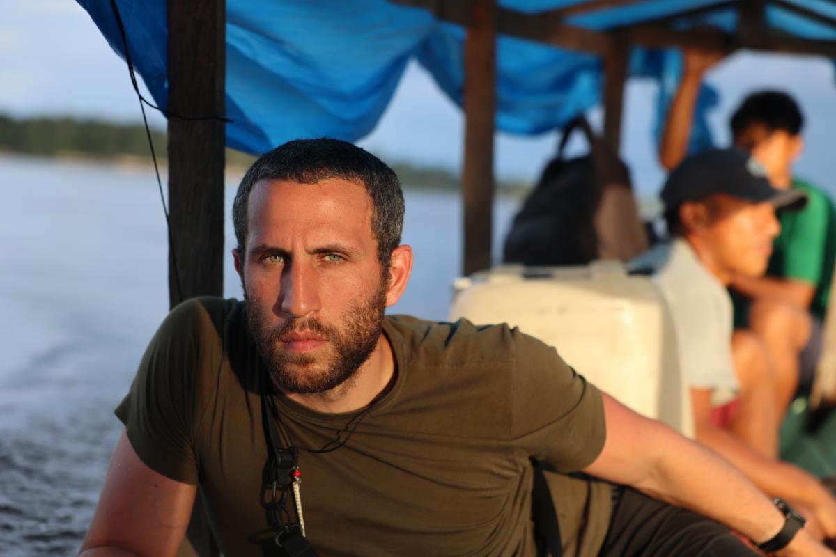 a man seated on a covered river boat with water and treeline visible in the background