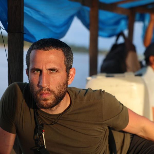 a man seated on a covered river boat with water and treeline visible in the background