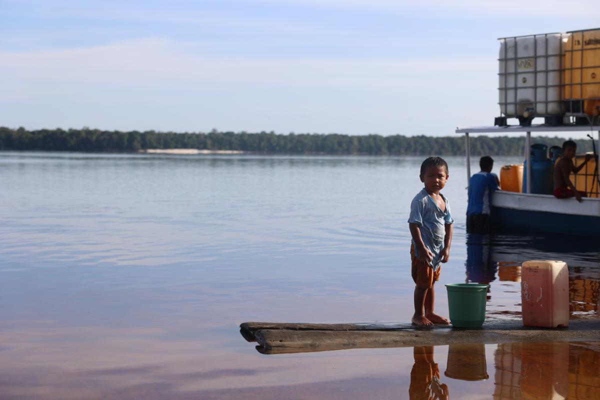 young child standing on a riverside jetty next to a moored boat and water containers, with a wide river and forested far bank in the background