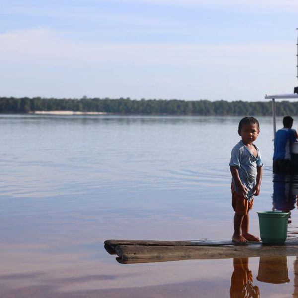 young child standing on a riverside jetty next to a moored boat and water containers, with a wide river and forested far bank in the background