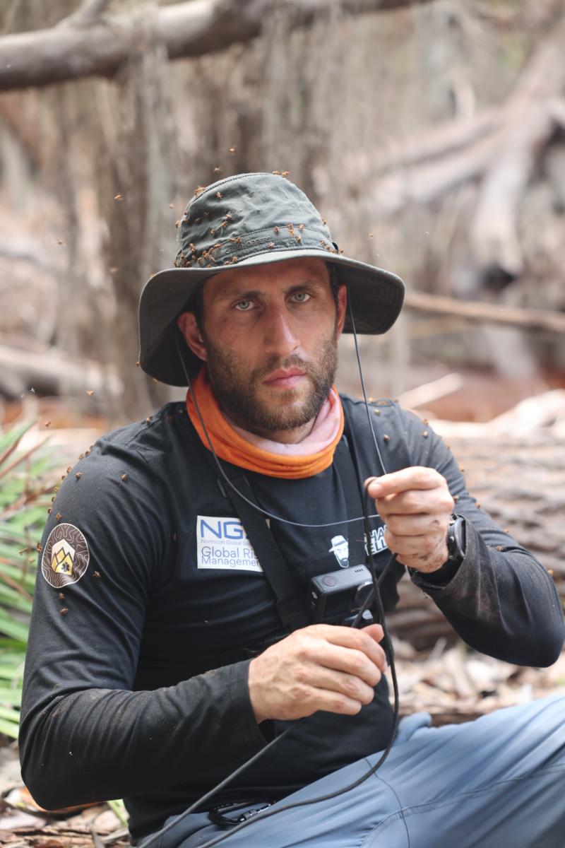 a man covered in insects sitting in a wooded outdoor setting, holding a camera and wearing branded field gear