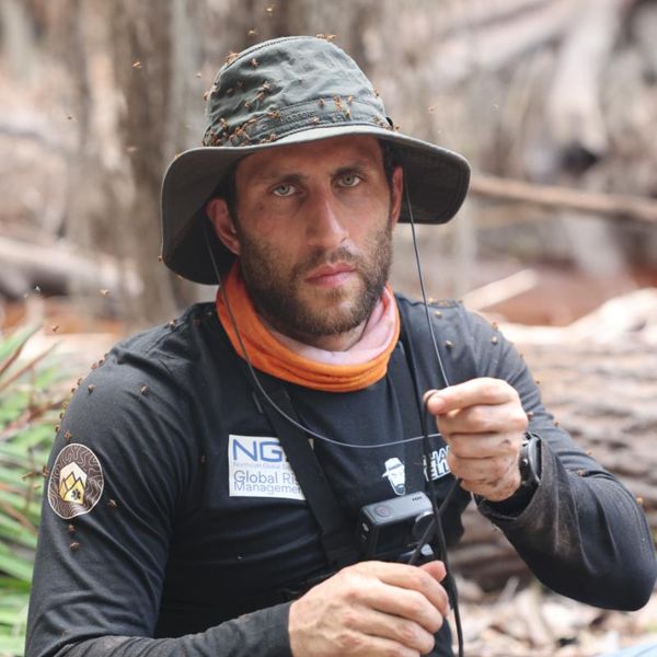 a man covered in insects sitting in a wooded outdoor setting, holding a camera and wearing branded field gear