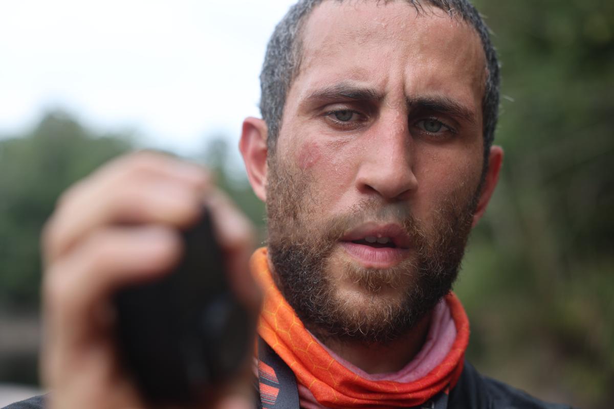 close-up portrait of a bearded man in an orange neck gaiter outdoors, with blurred greenery in the background