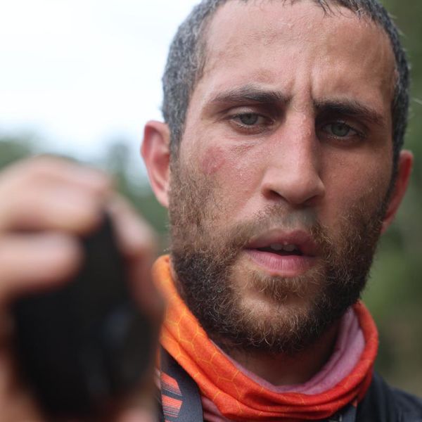 close-up portrait of a bearded man in an orange neck gaiter outdoors, with blurred greenery in the background