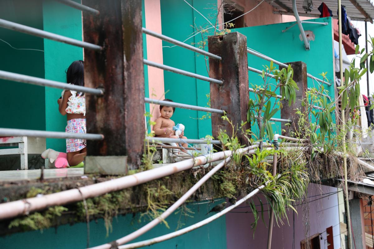 two young children on a weathered balcony of a colourful building, with metal railings and trailing plants