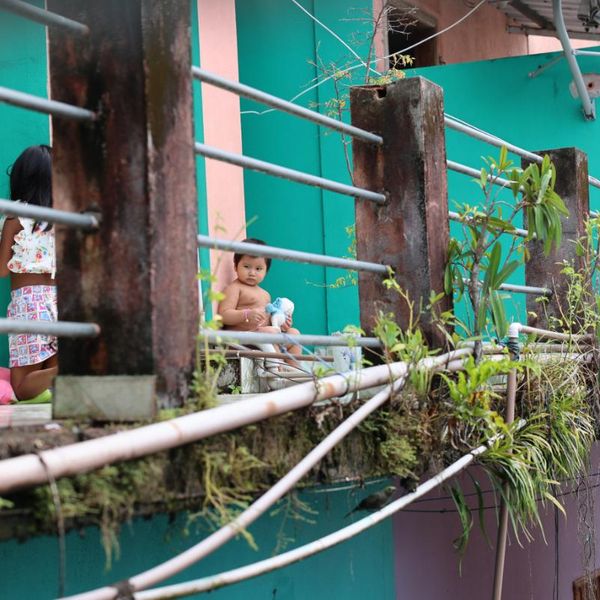two young children on a weathered balcony of a colourful building, with metal railings and trailing plants