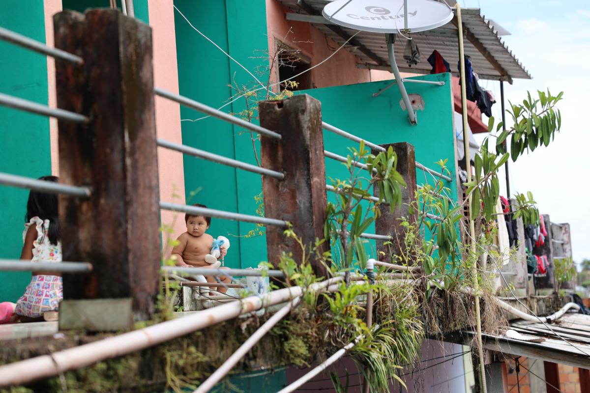 a toddler sitting on a railed walkway beside colourful stilt buildings, with laundry drying and a satellite dish visible nearby
