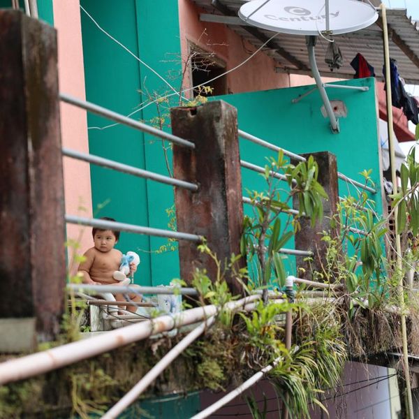 a toddler sitting on a railed walkway beside colourful stilt buildings, with laundry drying and a satellite dish visible nearby