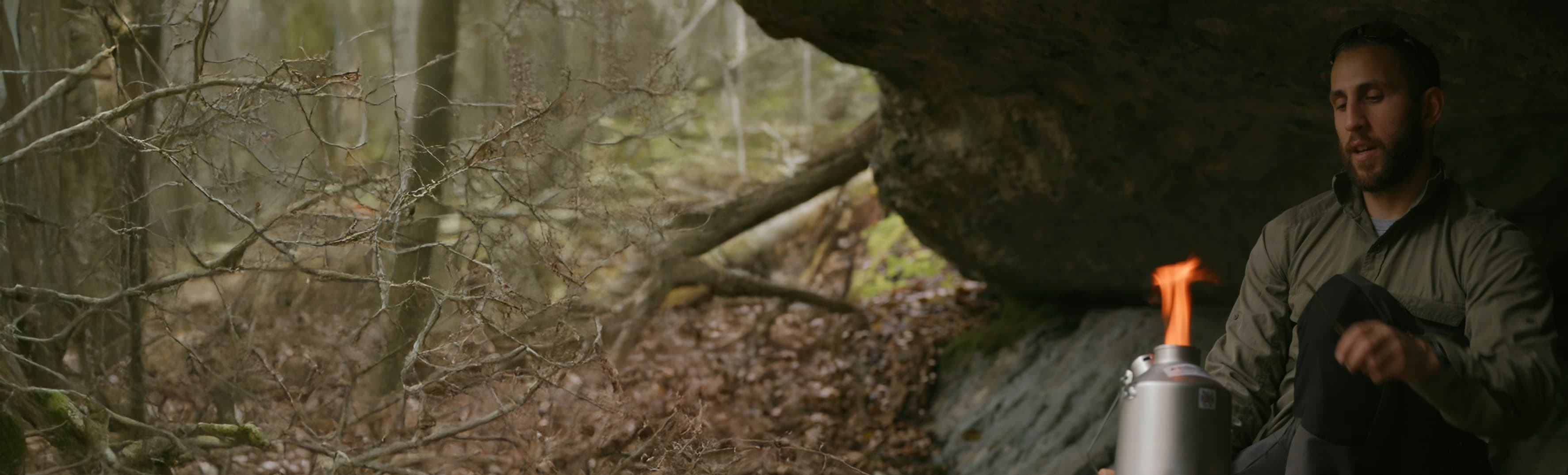 a man sheltering under a rock outcrop in a leafless forest, using a small backpacking stove with a visible flame
