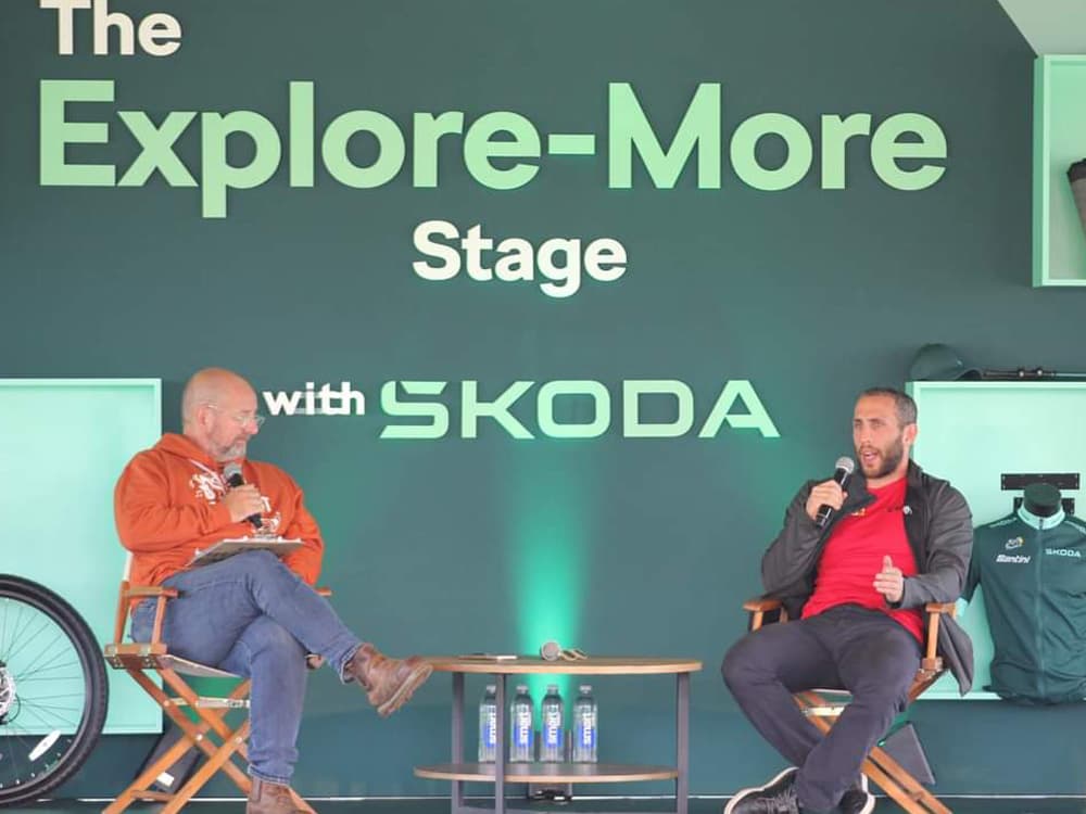 two speakers on stage at a branded Skoda Explore-More Stage event, seated on director's chairs with a cycling jersey display and bicycle wheel visible in the background