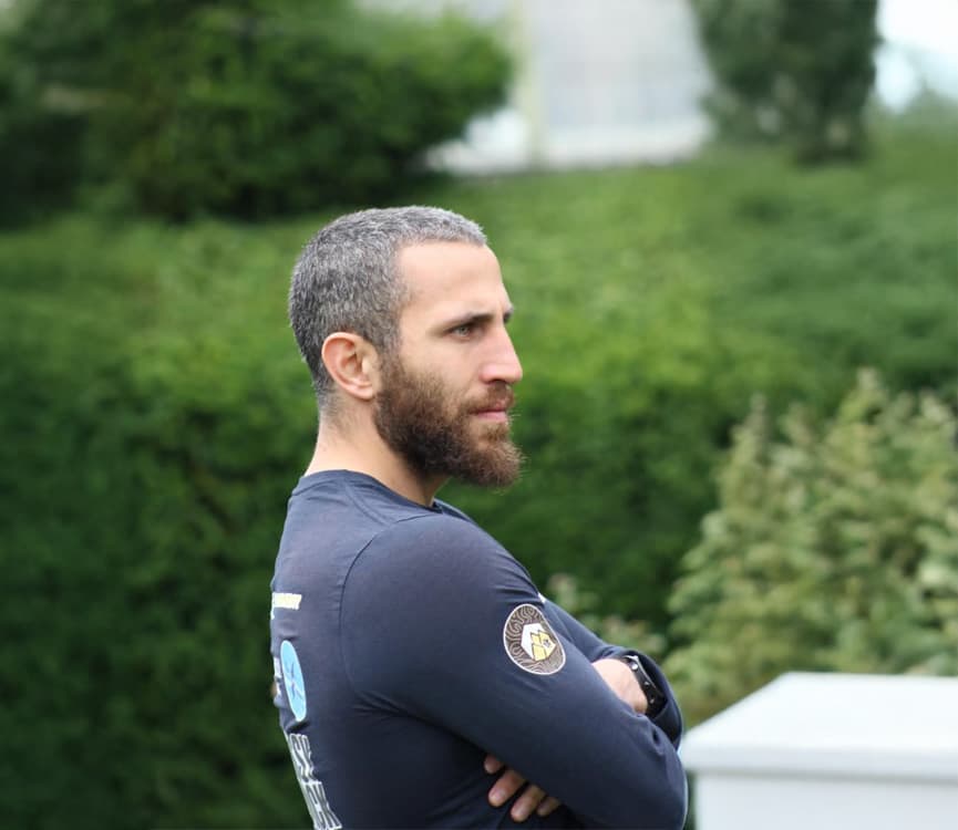 profile portrait of a bearded man with arms crossed outdoors, wearing a branded dark long-sleeve top